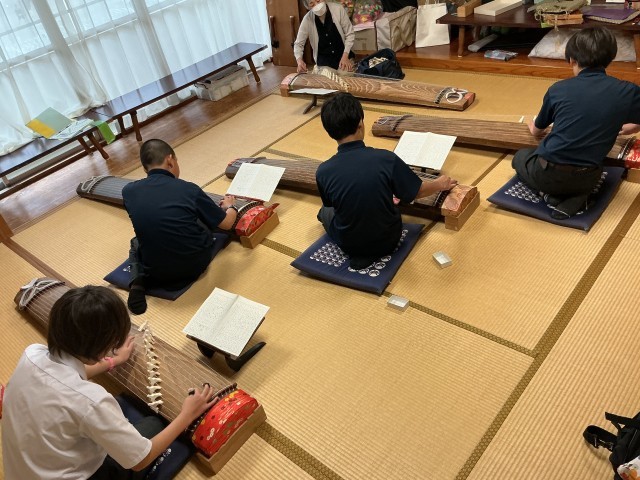1年生4名が和室に座り、前に箏の先生が座っている。全員で箏を弾き、稽古しているところ。