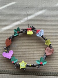 部員手作りのクリスマスリース。細い木の枝を束ねたものをつなげて輪っかにして、折り紙で作った飾りがついている。一番上には鈴、そこから時計回りににサンタクロース、大きい星、トナカイ、ヒイラギ、小さい星、大きい星、ヒイラギ、ハート、小さい星、トナカイ、ヒイラギが飾られている。