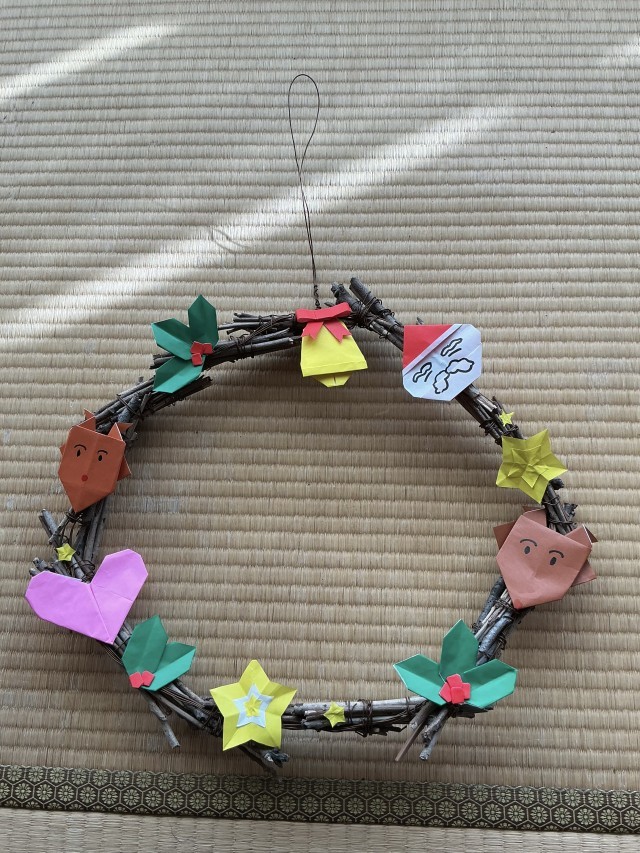 部員手作りのクリスマスリース。細い木の枝を束ねたものをつなげて輪っかにして、折り紙で作った飾りがついている。一番上には鈴、そこから時計回りににサンタクロース、大きい星、トナカイ、ヒイラギ、小さい星、大きい星、ヒイラギ、ハート、小さい星、トナカイ、ヒイラギが飾られている。