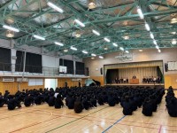 着任式が行われている様子を体育館後方から撮ったもの。生徒は座り、12名の着任職員が壇上の椅子に座り、演題で校長が話している。