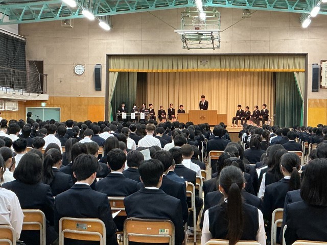   体育館で生徒総会が行われている。壇上に立つ生徒会長が、左下に立つ代表者からの質問に会長が回答している。